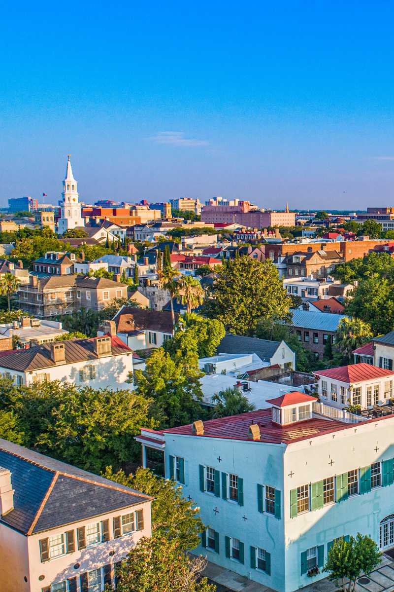 Michelle Almeyda-Wiedemuth An elevated cityscape view of Charleston's historic district, featuring a prominent white church steeple rising above traditional rooftops under a bright sky.